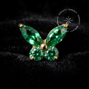 Piercing Labret Topo Borboleta Cravejada com Zircônia Verde em Titânio Revestido de Ouro 18k, repousando suavemente sobre um tecido de cetim branco de alta opulência, fotografado em alta definição com foco nos detalhes geométricos de sua estrutura.