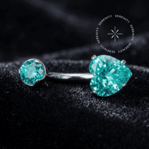 Piercing de Umbigo Titânio repousando suavemente sobre um tecido de cetim branco de alta opulência, destacando o polimento espelhado da joia e suas exclusivas gemas cravejadas na cor verde paraíba.