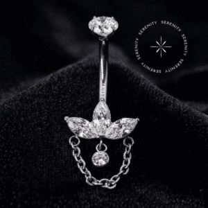 Piercing umbigo titânio com design composto por zircônias em lapidação navete e pingente de correntes delicadas, repousando suavemente sobre um tecido de cetim branco de alta opulência. O acabamento polido do metal reflete a iluminação requintada do ambiente.