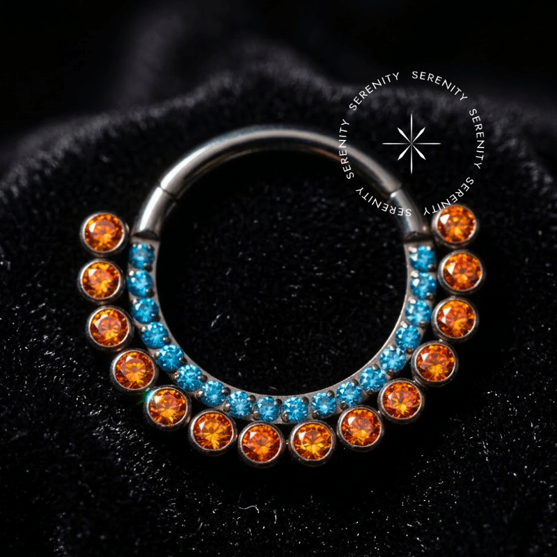 Argola Click Titânio com design frontal cravejado intercalando Zircônias Premium nas cores azul e laranja, fecho clicker em dobradiça invisível, ideal para perfurações de Daith e Septo.
