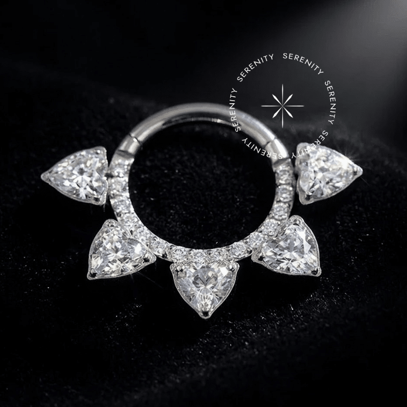 Piercing septo cravejado luxo com design em arco apresentando cinco pedras de Zircônia Premium em formato de coração, mecanismo de encaixe clicker e acabamento polido, ideal para Daith e Septo em setups de alta perfuração. Título: piercing-septo-cravejado-luxo-coracoes-tn132