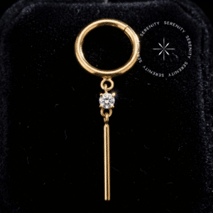 Argola clicker titânio dourado com pingente em barra vertical cravejado de Zircônias Premium translúcidas, repousando sobre fundo liso, da exclusiva marca Serenity.
