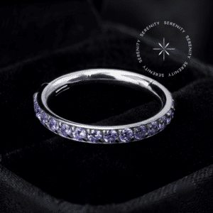 Piercing titânio grau implante argola clicker cravejada violeta repousando suavemente sobre um tecido de cetim branco de alta opulência, refletindo luminosidade intensa.