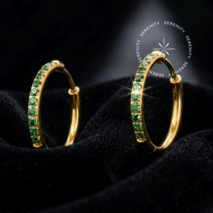 Fotografia macro do Brinco Argola Cravejada em Titânio Revestido de Ouro 18k com zircônias verdes, repousando suavemente sobre um tecido de cetim branco de alta opulência.