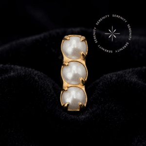 piercing titânio dourado em formato de barra com três pérolas sintéticas, exibindo polimento espelhado em um cenário de alto contraste e iluminação dramática.
