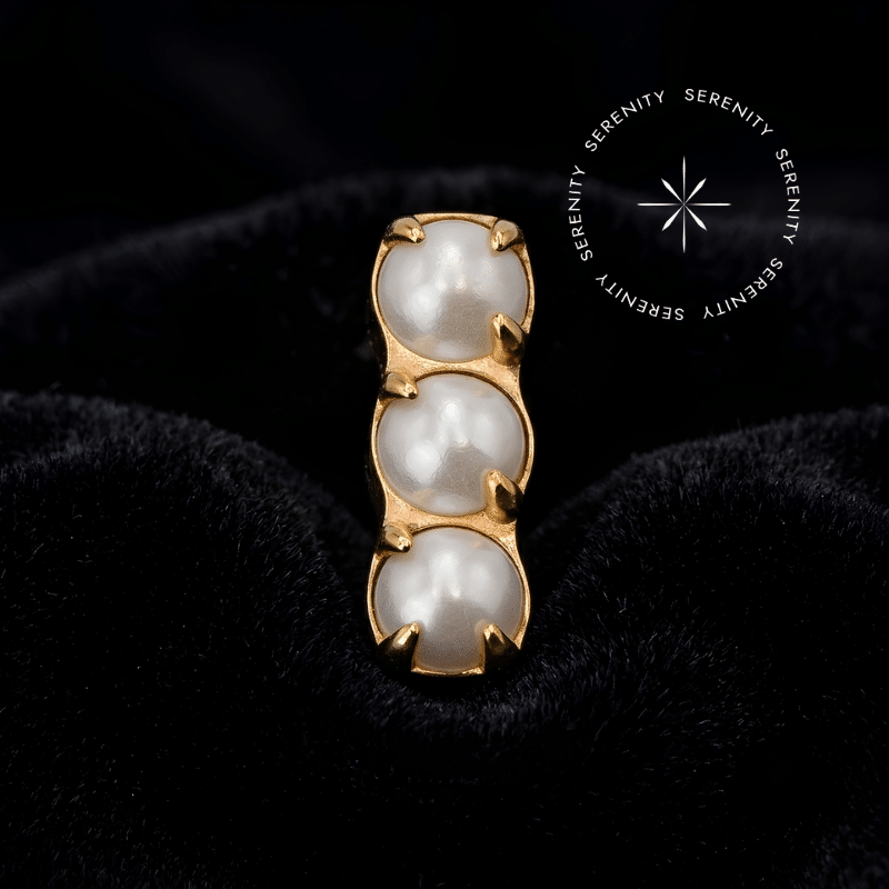 piercing titânio dourado em formato de barra com três pérolas sintéticas, exibindo polimento espelhado em um cenário de alto contraste e iluminação dramática.