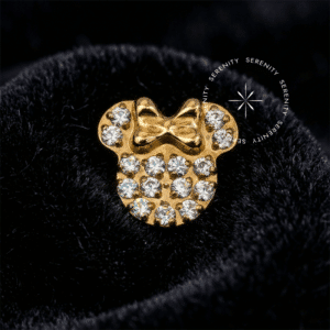 Piercing Titânio Dourado com topo esculpido no formato da silhueta da Minnie Mouse, enriquecido com um detalhe de laço, totalmente cravejado em pavê com Zircônias Premium Radiantes. Pino rosqueável inferior de 0.9 mm para fixação em labrets, exibido sobre fundo branco ressaltando a captação de luz e o luxo.