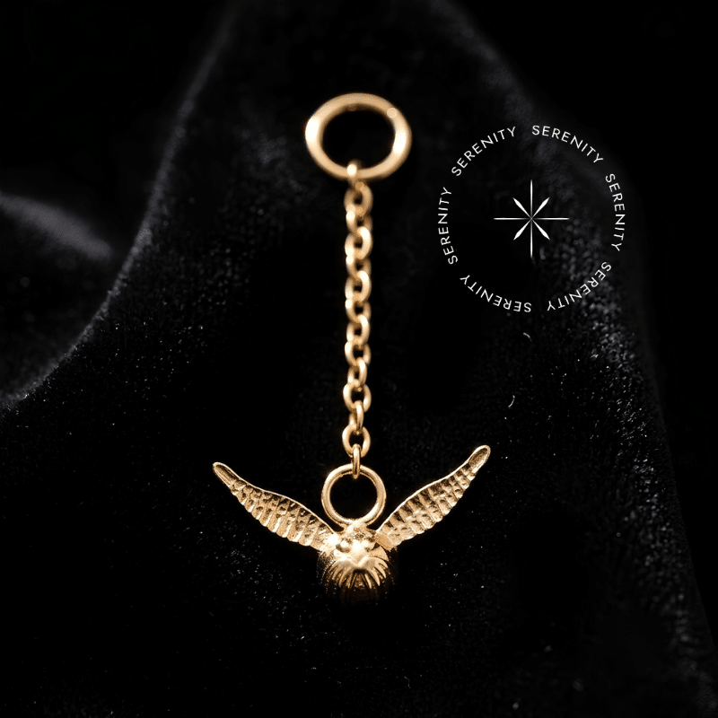 Corrente Piercing Titânio com pingente lúdico em formato de Pomo e asas abertas, textura polida e espelhada em ouro 18k, argolas de encaixe nas extremidades para interligação em projetos de perfuração e alta joalheria piercing.