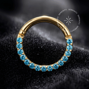 Argola clicker titânio dourado com cravação frontal pavé de zircônias premium na cor azul imperial intensa, sistema de fecho articulado suíço de alta precisão usinado em titânio grau implante revestido de ouro 18k, desenhada para perfurações de septo e daith.