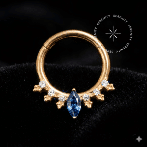 argola daith titânio em Titânio Grau Implante ASTM F136 com acabamento PVD em Ouro 18k, exibindo design com cravação frontal de pedra central Zircônia Premium corte Navete em tom Azul Imperial emoldurada por coroa simétrica de esferas polidas e micro zircônias claras, com sistema de abertura articulada clicker de precisão ideal para adorno corporal de luxo no daith e septo.