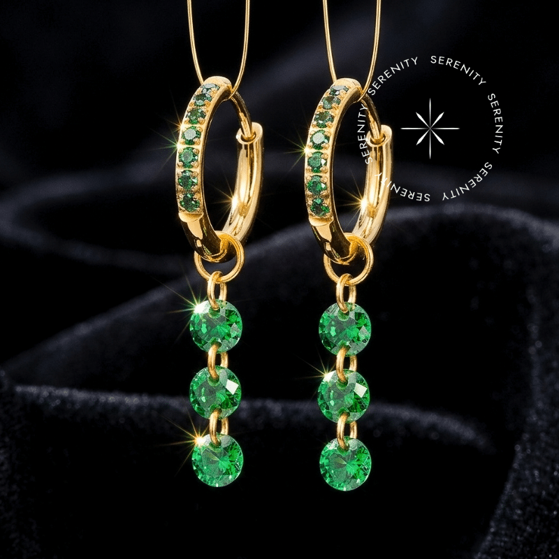 Brinco Argola Titânio dourado com três pingentes móveis cravejados com zircônias esmeralda, sistema de encaixe lateral para lóbulo repousando em ambiente de alto luxo.
