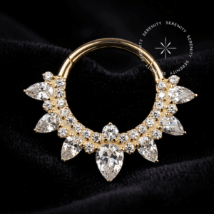Fotografia em modo macro de uma argola clicker titânio dourado cravejada com zircônias translúcidas em formato de gota e redondas, repousando suavemente sobre um tecido de cetim branco de alta opulência.