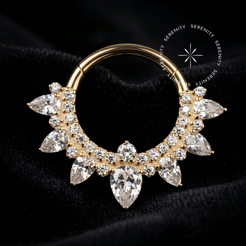 Fotografia em modo macro de uma argola clicker titânio dourado cravejada com zircônias translúcidas em formato de gota e redondas, repousando suavemente sobre um tecido de cetim branco de alta opulência.