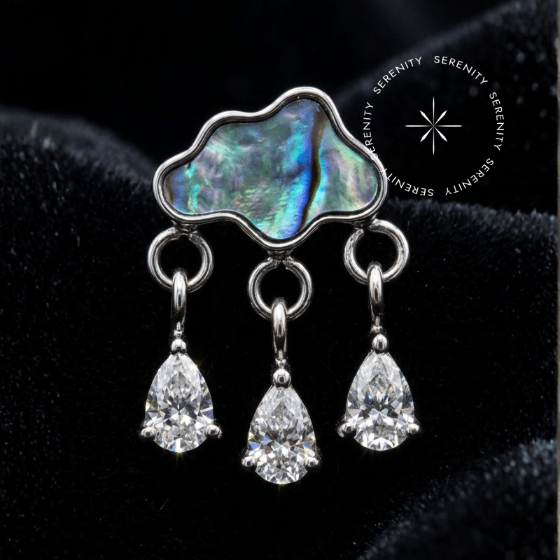 piercing titânio grau implante com design de nuvem adornado com pedra natural abalone e três pingentes em zircônia translúcida repousando suavemente sobre um tecido de cetim branco de alta opulência.