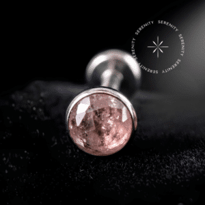 Piercing Topo Titânio repousando suavemente sobre um tecido de veludo de seda preto de alta opulência, exibindo sua cravação protetora ao redor de uma pedra natural rosa de 4mm.