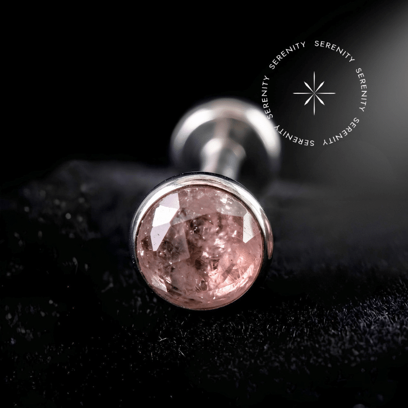Piercing Topo Titânio repousando suavemente sobre um tecido de veludo de seda preto de alta opulência, exibindo sua cravação protetora ao redor de uma pedra natural rosa de 4mm.