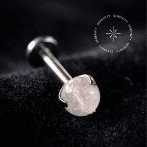 Piercing Topo Titânio repousando suavemente sobre um tecido de cetim branco de alta opulência, exibindo sua cravação em garras ao redor de uma esfera de pedra natural strawberry de 4mm.