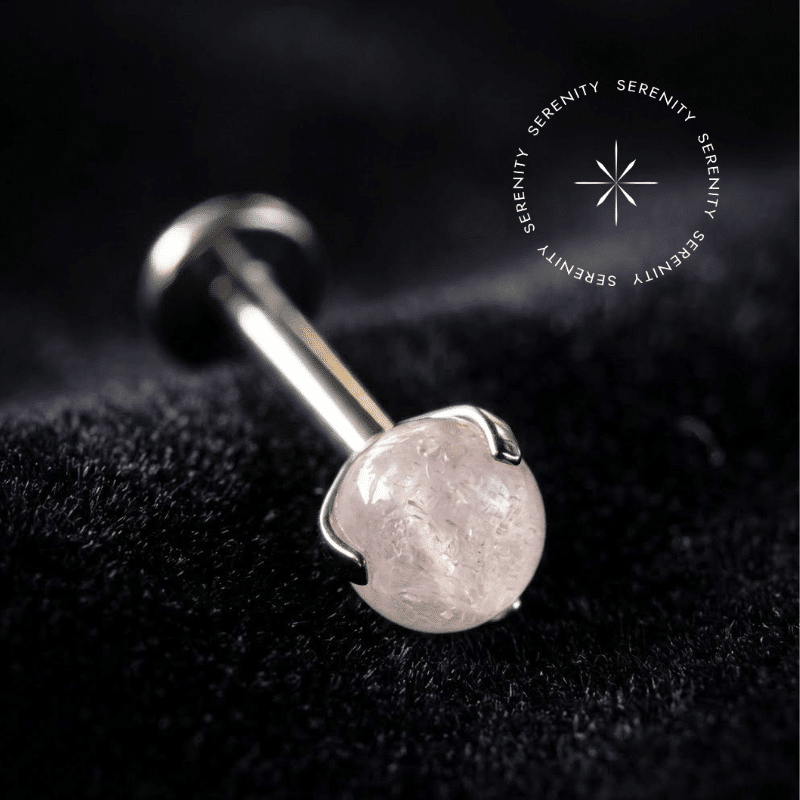 Piercing Topo Titânio repousando suavemente sobre um tecido de cetim branco de alta opulência, exibindo sua cravação em garras ao redor de uma esfera de pedra natural strawberry de 4mm.
