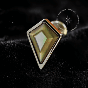Piercing Topo Titânio repousando suavemente sobre um tecido de cetim preto de alta opulência, evidenciando seu expressivo topo de 9mm em formato de losango preenchido com pedra natural abalone multicolorida.