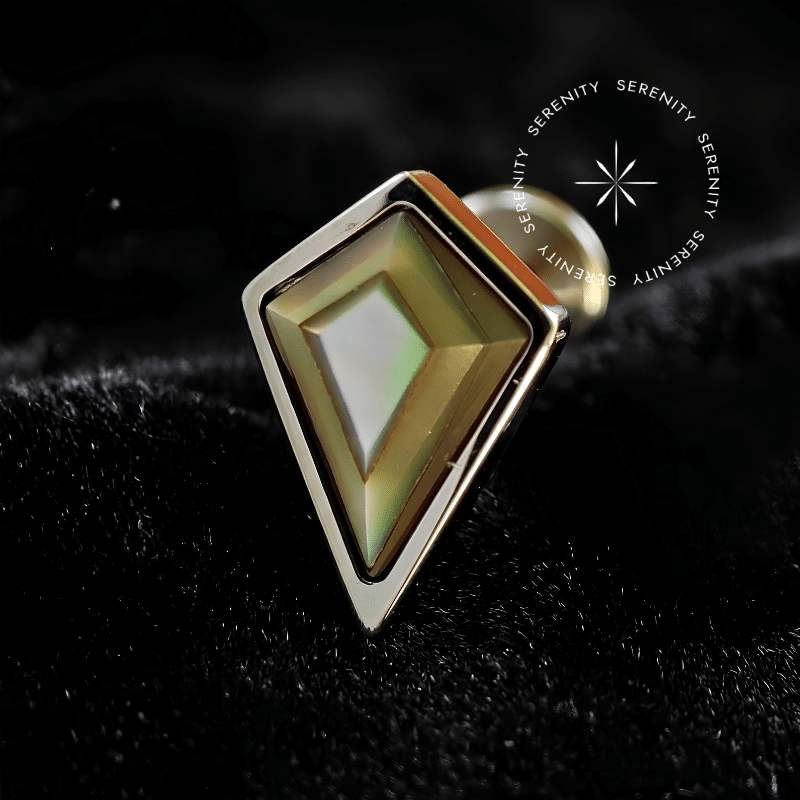 Piercing Topo Titânio repousando suavemente sobre um tecido de cetim preto de alta opulência, evidenciando seu expressivo topo de 9mm em formato de losango preenchido com pedra natural abalone multicolorida.