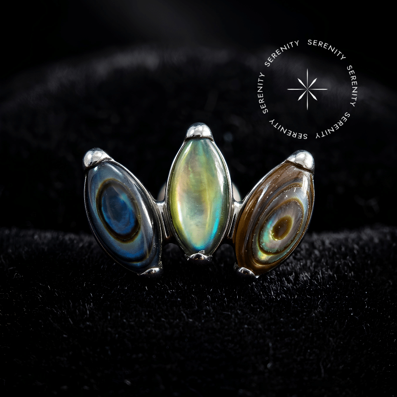 piercing titânio grau implante com design cluster triplo adornado por autênticas pedras naturais abalone repousando suavemente sobre um tecido de cetim branco de alta opulência.