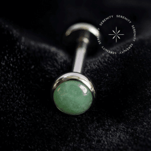 Piercing Topo Titânio repousando suavemente sobre um tecido de cetim preto de alta opulência, exibindo sua pedra natural Jade Malásia de 4mm perfeitamente protegida por uma moldura lisa e polida.