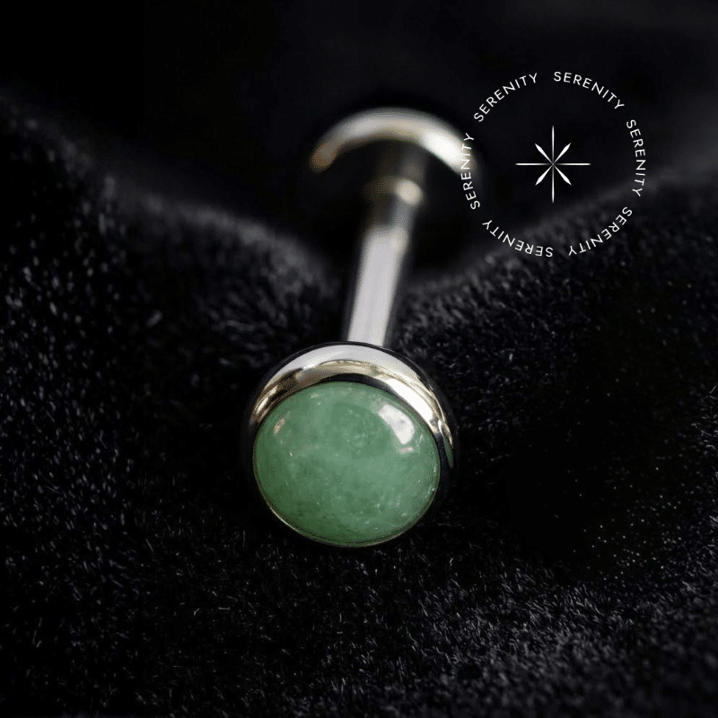Piercing Topo Titânio repousando suavemente sobre um tecido de cetim preto de alta opulência, exibindo sua pedra natural Jade Malásia de 4mm perfeitamente protegida por uma moldura lisa e polida.