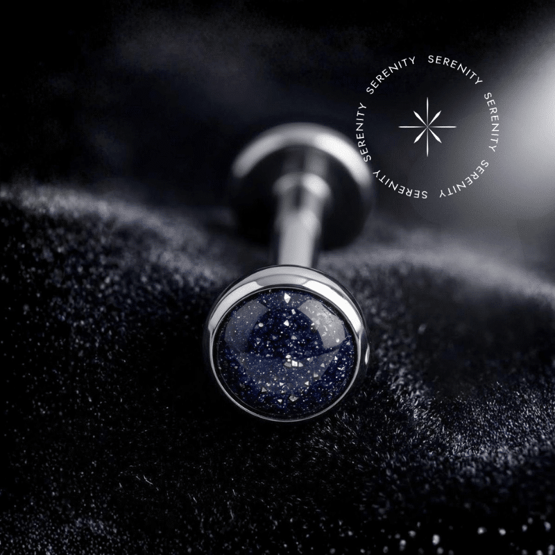 Piercing Topo Titânio repousando suavemente sobre um tecido de cetim preto de alta opulência, exibindo sua pedra natural ônix com pontos brilhantes perfeitamente protegida por uma moldura lisa e polida.