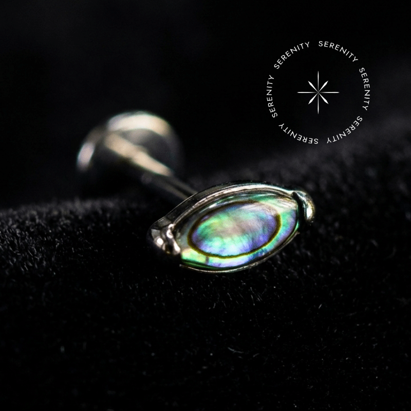 piercing titânio grau implante com topo oval de 4mm adornado por autêntica pedra natural abalone, repousando suavemente sobre um tecido de cetim branco de alta opulência.