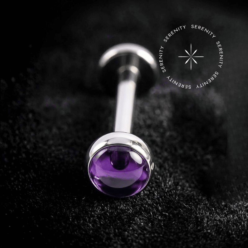 Piercing Topo Titânio repousando suavemente sobre um tecido de cetim preto de alta opulência, exibindo sua pedra natural ametista de 4mm perfeitamente protegida por uma moldura lisa e polida.