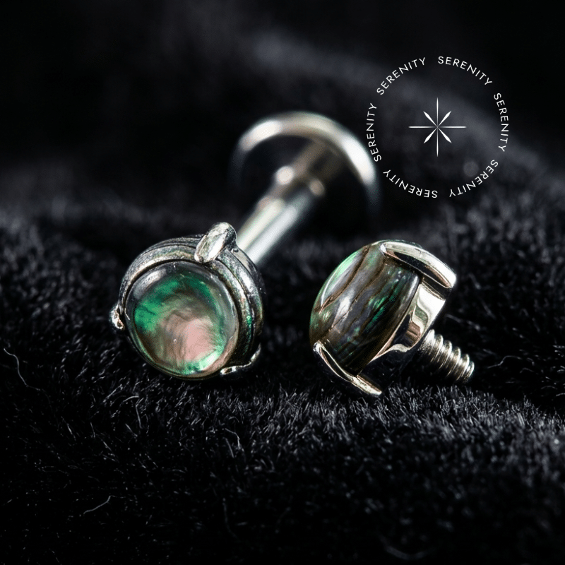 piercing titânio grau implante com topo circular minimalista de 3mm adornado por autêntica pedra natural abalone, repousando suavemente sobre um tecido de cetim branco de alta opulência.
