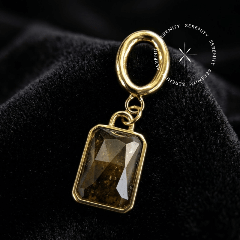 Piercing Pingente de Orelha Titânio com Pedra Natural Marrom em lapidação retangular repousando suavemente sobre um tecido de cetim preto de alta opulência, destacando o banho dourado.