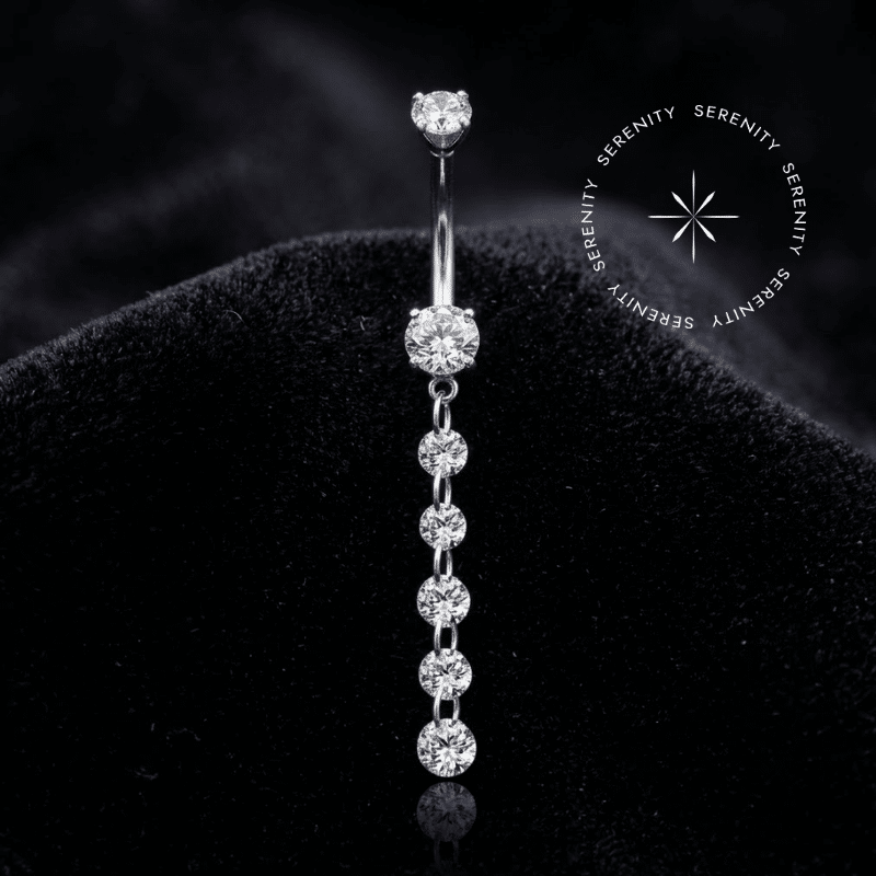 Piercing Umbigo Pingente Titânio repousando suavemente sobre um tecido de cetim branco de alta opulência, evidenciando o brilho profundo de suas zircônias translúcidas e o acabamento espelhado impecável do titânio natural platinado.