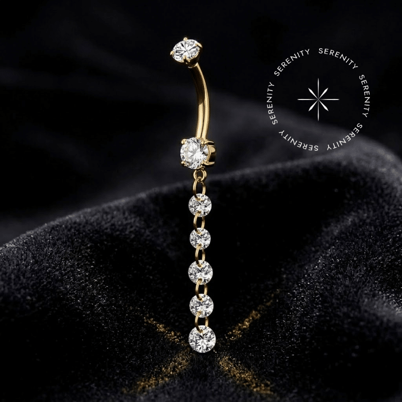 Piercing Umbigo Pingente Titânio repousando suavemente sobre um tecido de cetim preto de alta opulência, evidenciando o brilho profundo de suas zircônias e o acabamento espelhado do revestimento em ouro 18k.