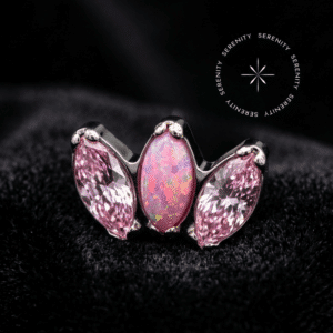piercing titânio grau implante com design tríade adornado por opala rosa central e zircônias laterais, repousando suavemente sobre um tecido de cetim branco de alta opulência.