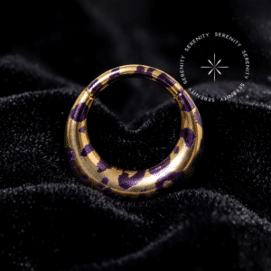 Piercing Argola Septo Titânio repousando suavemente sobre um veludo de seda preto de alta opulência, destacando o formato oval abaulado e a estampa de oncinha roxa sobre o dourado.