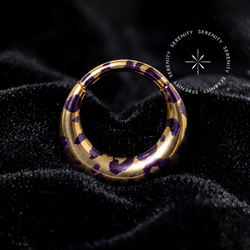 Piercing Argola Septo Titânio repousando suavemente sobre um veludo de seda preto de alta opulência, destacando o formato oval abaulado e a estampa de oncinha roxa sobre o dourado.