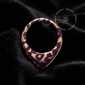 Piercing Argola Septo Titânio repousando suavemente sobre um tecido de cetim preto de alta opulência, destacando o formato em V e a estampa de oncinha roxa com detalhes dourados.
