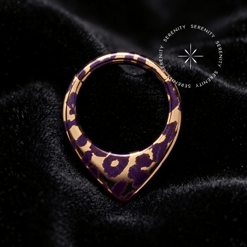 Piercing Argola Septo Titânio repousando suavemente sobre um tecido de cetim preto de alta opulência, destacando o formato em V e a estampa de oncinha roxa com detalhes dourados.
