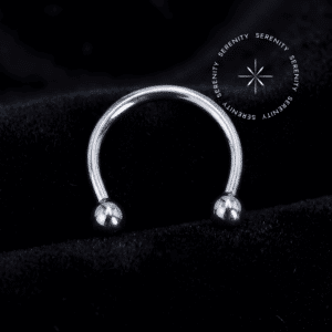 Fotografia macro do Piercing Ferradura Titânio com sistema push pin, de polimento impecável, repousando suavemente sobre um tecido de cetim branco de alta opulência.