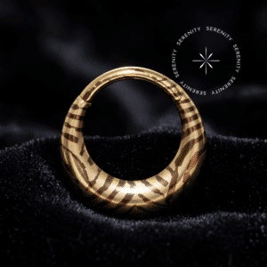Piercing Argola Septo Titânio repousando suavemente sobre um tecido de cetim preto de alta opulência, destacando o formato oval abaulado e a estampa listrada em tons dourados e escuros.