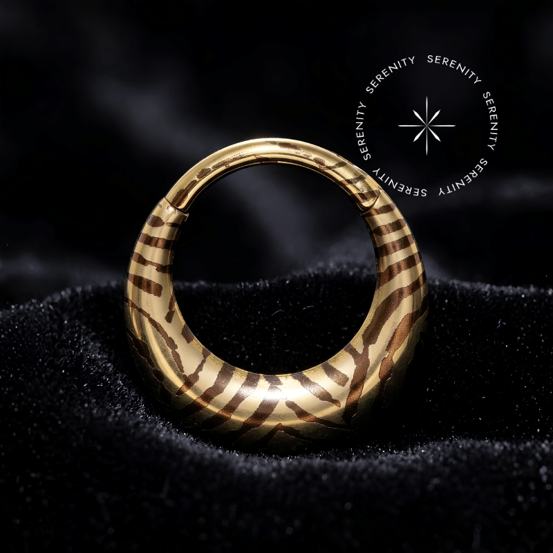Piercing Argola Septo Titânio repousando suavemente sobre um tecido de cetim preto de alta opulência, destacando o formato oval abaulado e a estampa listrada em tons dourados e escuros.
