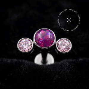 piercing titânio grau implante com design cluster adornado por opala magenta central e zircônias rosas laterais, repousando suavemente sobre um tecido de cetim branco de alta opulência.