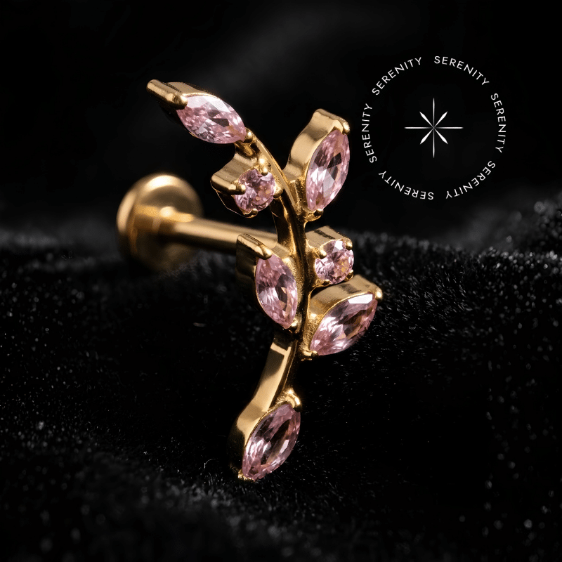 Piercing Topo de Orelha Titânio repousando suavemente sobre um tecido de veludo de seda preto de alta opulência, destacando o formato de ramo dourado com zircônias cor rosa e o brilho do metal nobre.