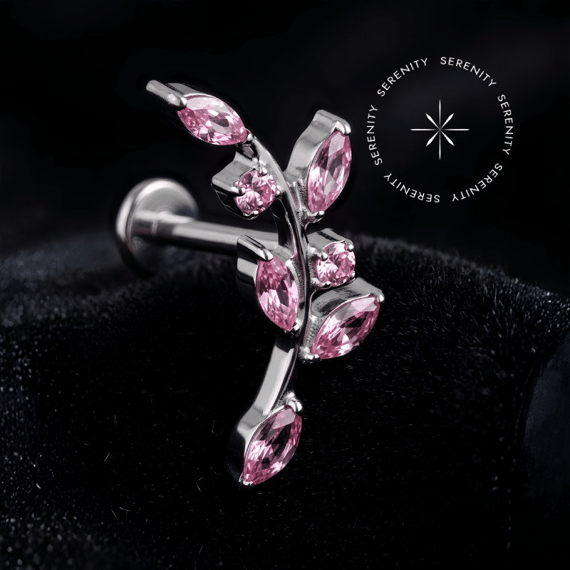 Piercing Topo de Orelha Titânio repousando suavemente sobre um tecido de cetim preto de alta opulência, destacando o formato de ramo com zircônias cor rosa e o brilho do metal.