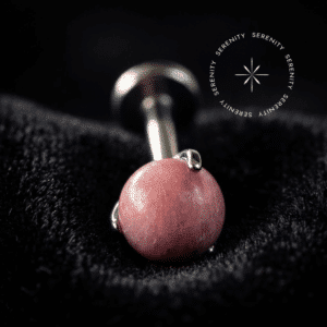 piercing topo pedra natural rhodonite repousando suavemente sobre um tecido de cetim branco de alta opulência, destacando o polimento profundo do titânio e os tons rosados terrosos da esfera orgânica.