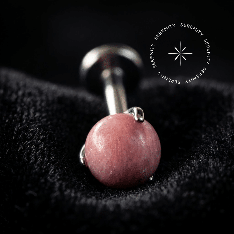 piercing topo pedra natural rhodonite repousando suavemente sobre um tecido de cetim branco de alta opulência, destacando o polimento profundo do titânio e os tons rosados terrosos da esfera orgânica.