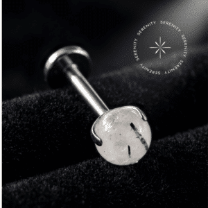 piercing topo pedra natural black rutile quartz repousando suavemente sobre um tecido de cetim branco de alta opulência, destacando o polimento profundo do titânio e as inclusões minerais da esfera orgânica.