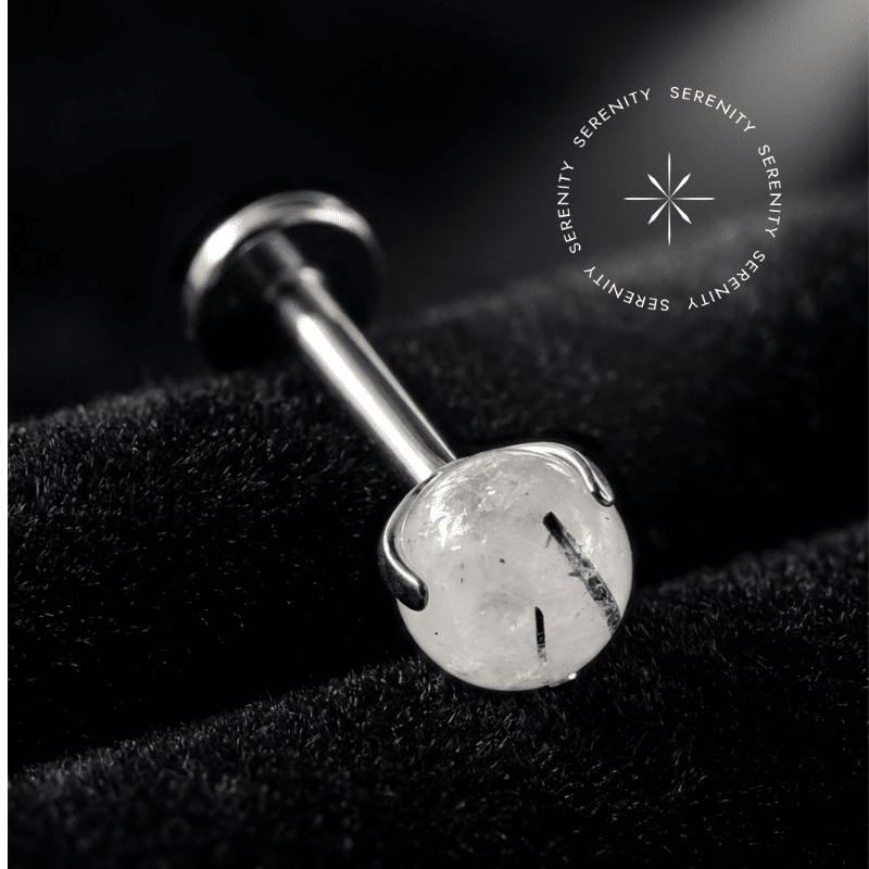 piercing topo pedra natural black rutile quartz repousando suavemente sobre um tecido de cetim branco de alta opulência, destacando o polimento profundo do titânio e as inclusões minerais da esfera orgânica.