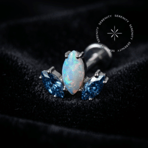 Topo Trio Cravejado em Titânio repousando suavemente sobre um tecido de cetim branco de alta opulência, destacando a iridescência da opala central e o azul vibrante das zircônias laterais.